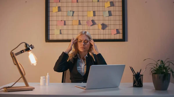 Mujer de negocios que sufre de dolor de cabeza cerca de píldoras y aerosol nasal en la oficina por la noche - foto de stock