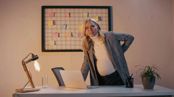 Femme d'affaires enceinte souriante debout près de l'ordinateur portable sur la table de bureau — Photo de stock