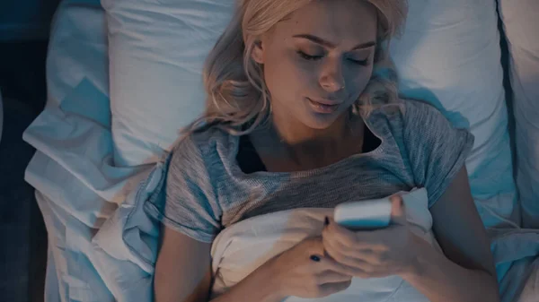 Вид сверху на женщину, использующую смартфон, лежа на кровати — стоковое фото