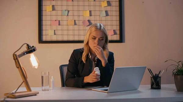 Donna d'affari nervosa che tiene il vaso con pillole vicino a computer portatile in ufficio — Stock Photo