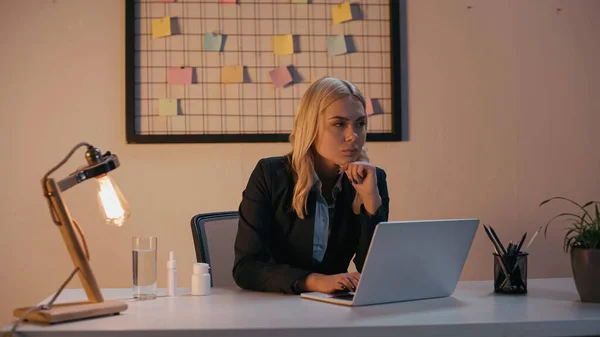 Femme d'affaires regardant loin près de l'ordinateur portable et des pilules sur la table de bureau — Photo de stock