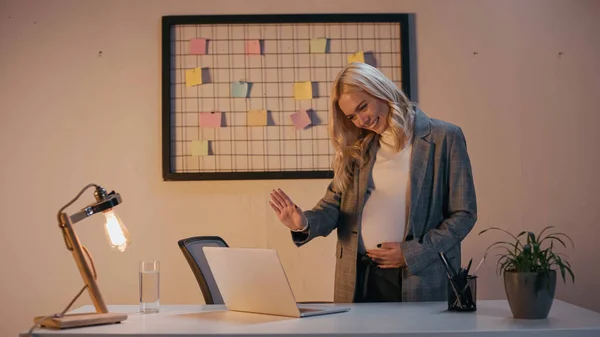 Smiling pregnant businesswoman waving hand at laptop in office — Stock Photo