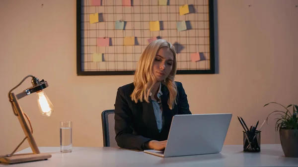 Businesswoman using laptop near lamp and glass of water in office in evening — Stock Photo