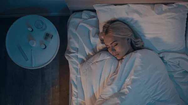 Vista dall'alto della donna che dorme sul letto vicino a pillole e termometro elettronico in camera da letto — Foto stock