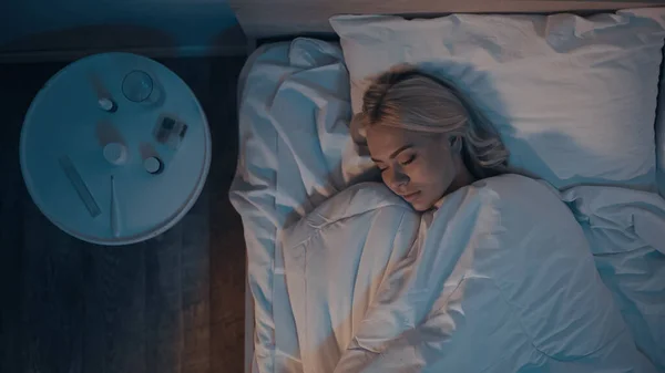 Vista dall'alto della donna che dorme sul letto vicino a pillole e termometro elettronico in camera da letto — Stock Photo