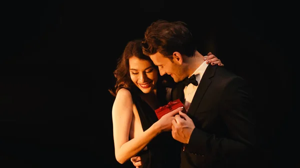 Heureuse jeune femme recevant boîte cadeau et câlin homme joyeux en costume isolé sur noir — Photo de stock