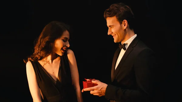 Excited young woman receiving gift box from man in suit isolated on black — Stock Photo