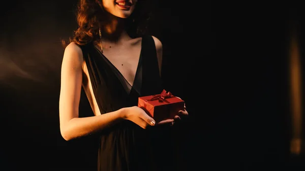 Vista recortada de la joven feliz sosteniendo caja de regalo roja en negro - foto de stock