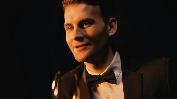Sonriente joven en traje con corbata de lazo sosteniendo vidrio aislado en negro - foto de stock