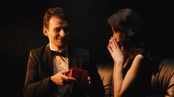 Happy young man in suit with bow tie giving present to stunned woman isolated on black — Stock Photo