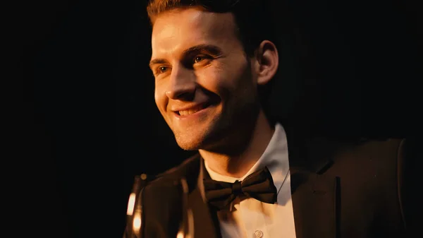 Hombre joven feliz en traje con corbata de lazo sosteniendo vidrio aislado en negro - foto de stock