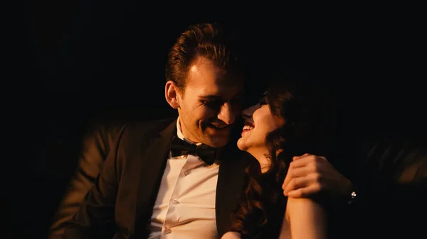 Smiling man in suit hugging and flirting with joyful girlfriend isolated on black — Stock Photo