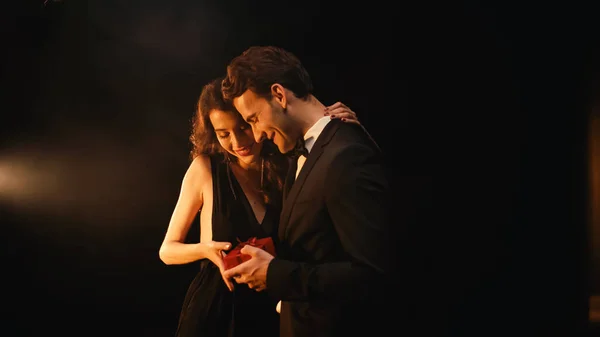 Hombre joven feliz en traje con pajarita dando regalo rojo a la mujer en vestido en negro - foto de stock