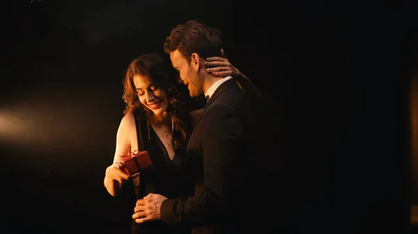 Giovane uomo sorridente in giacca e cravatta dare regalo rosso alla donna felice in abito nero — Foto stock