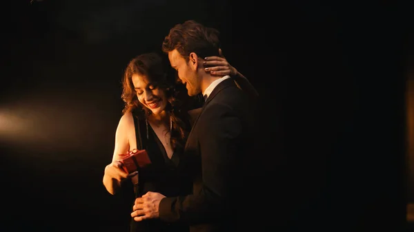 Sonriente joven en traje con pajarita dando regalo rojo a mujer feliz en vestido en negro - foto de stock