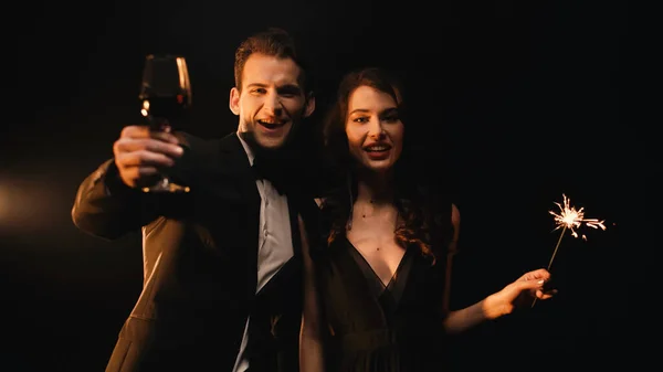 Allegro uomo in possesso di un bicchiere di vino rosso e donna con sparkler guardando la fotocamera sul nero — Foto stock