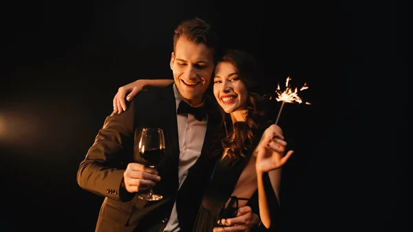 Woman with sparkler hugging happy man holding glass of red wine on black — Stock Photo