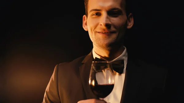 Joyeux jeune homme en costume avec noeud papillon tenant verre de vin rouge sur noir — Photo de stock