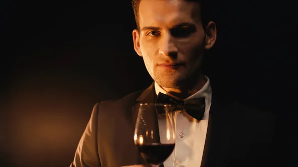 Confident man in suit with bow tie holding glass of red wine on black — Stock Photo