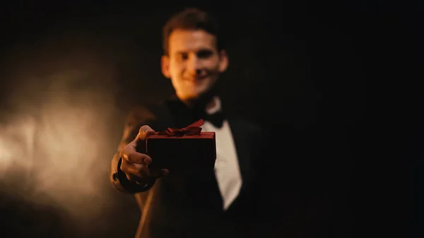 Hombre feliz y borroso en traje mostrando caja de regalo roja en negro - foto de stock