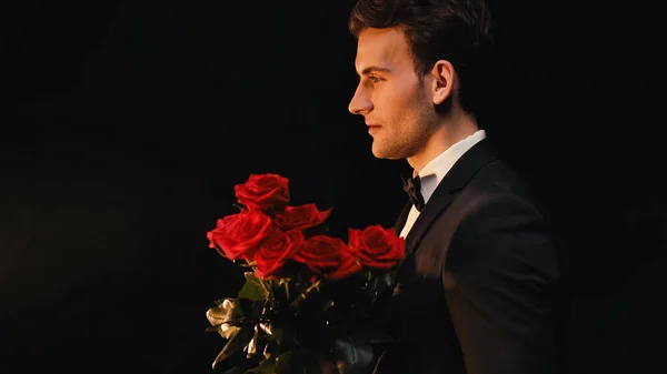 Vista lateral de un joven elegante sosteniendo rosas rojas aisladas en negro - foto de stock