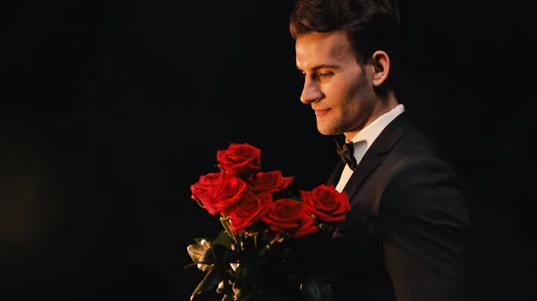 Joven elegante hombre sosteniendo rosas rojas aisladas en negro - foto de stock