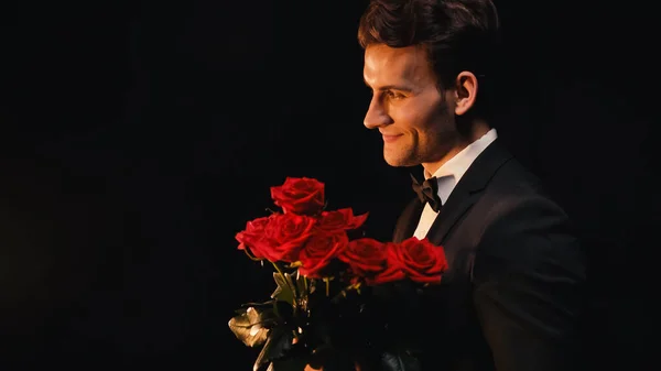 Vue latérale d'un jeune homme joyeux tenant des roses rouges isolées sur du noir — Photo de stock
