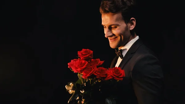 Щасливий молодий чоловік у костюмі тримає червоні троянди ізольовані на чорному — стокове фото