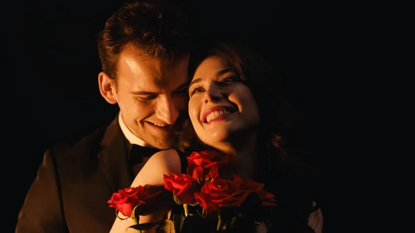 Fröhliche Frau erhält rote Rosen von Mann, der hinter isoliert auf schwarz steht — Stockfoto