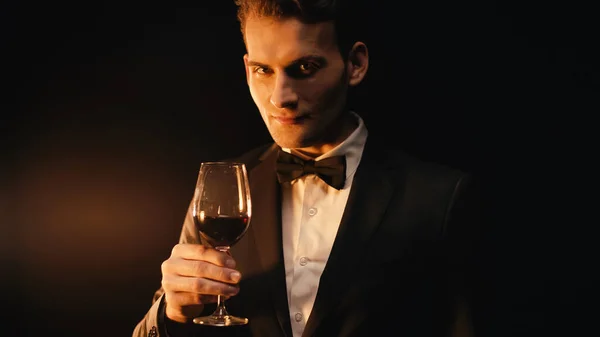 Elegant young man in suit with bow tie holding glass of wine on black — Stock Photo
