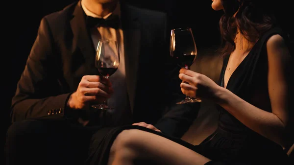 Vista recortada de la pareja sosteniendo vasos de vino tinto mientras está sentado en el sofá aislado en negro - foto de stock