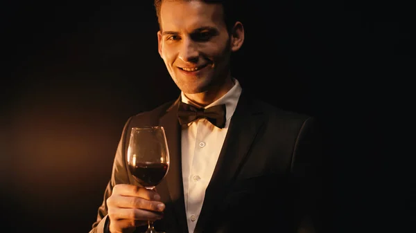 Fröhlicher junger Mann im Anzug mit Fliege, der ein Glas Wein auf schwarz hält — Stockfoto