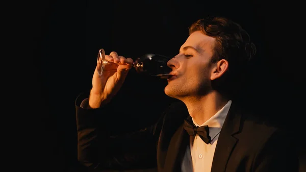 Elegante joven de traje con corbata de lazo sosteniendo vidrio y bebiendo vino aislado en negro - foto de stock