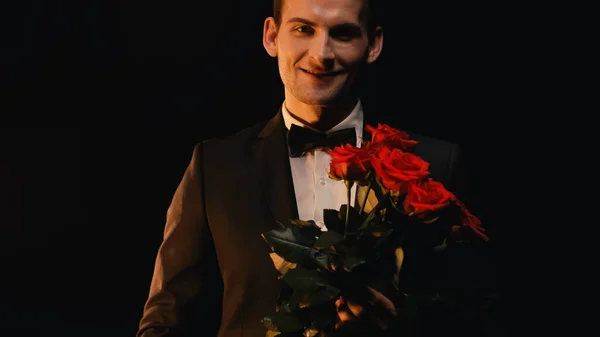 Feliz joven sosteniendo rosas rojas y sonriendo aislado en negro - foto de stock