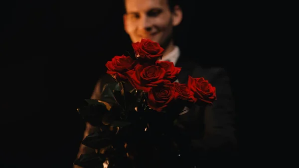 Hombre feliz y borroso sosteniendo rosas rojas aisladas en negro - foto de stock