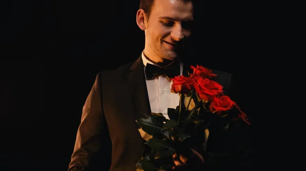 Joven feliz mirando rosas rojas y sonriendo aislado en negro - foto de stock