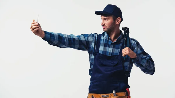 Arbeiter in Overalls und Mütze hält einen Rohrschlüssel in der Hand, während er ein Selfie auf weißem Grund macht — Stockfoto