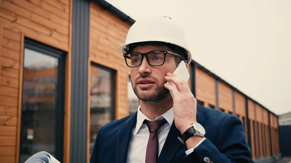 Arquiteto em hardhat falando no smartphone perto de prédio embaçado ao ar livre — Fotografia de Stock