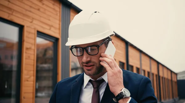 Ingeniero de traje hablando por teléfono inteligente cerca de un edificio borroso al aire libre - foto de stock