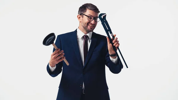 Hombre de negocios sonriente mirando la llave de la tubería y sosteniendo el émbolo aislado en blanco - foto de stock