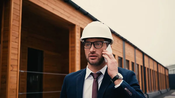 Engineer in white hardhat talking on mobile phone near building on urban street — Stock Photo