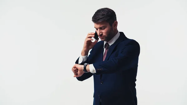 Manager in suit looking at wristwatch while talking on smartphone isolated on white — Stock Photo