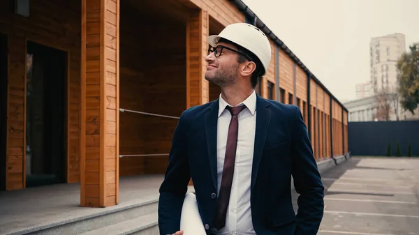 Side view of smiling engineer in hardhat holding blueprint and looking at building outdoors — Stock Photo