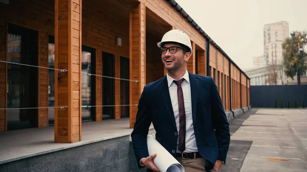 Joyeux architecte avec plan tenant la main dans la poche près du bâtiment à l'extérieur — Photo de stock