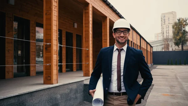 Позитивный инженер в защитном шлеме держит чертеж и смотрит на камеру возле здания на открытом воздухе — стоковое фото