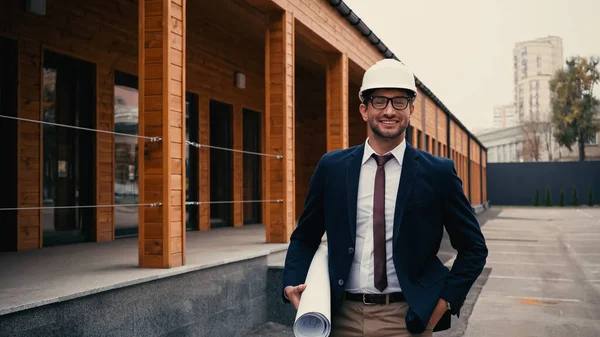 Ingegnere positivo in casco di sicurezza tenendo cianografia e guardando la fotocamera vicino edificio all'aperto — Foto stock