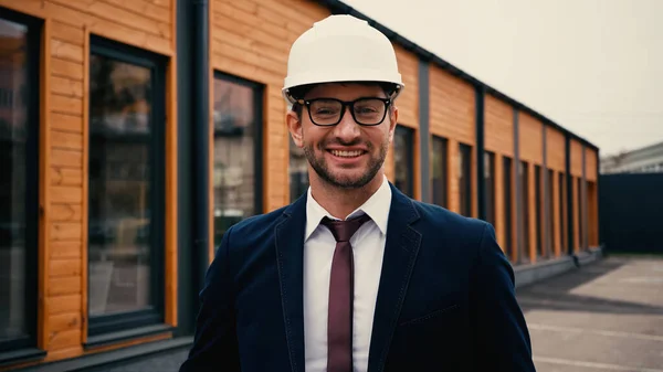 Lächelnder Ingenieur mit weißem Hut blickt in die Kamera in der Nähe von Gebäuden im Freien — Stockfoto