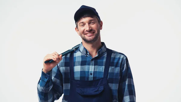 Glücklicher Arbeiter in Mütze und Overalls, der einen Rohrschlüssel auf weiß isoliert hält — Stockfoto