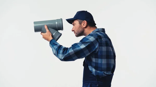 Seitenansicht des Heimwerkers in Overalls und Mütze, der durch ein auf Weiß isoliertes Kunststoffrohr blickt — Stockfoto