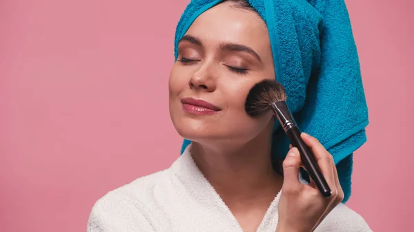 Mujer complacida con los ojos cerrados aplicando polvo facial con cepillo cosmético aislado en rosa - foto de stock